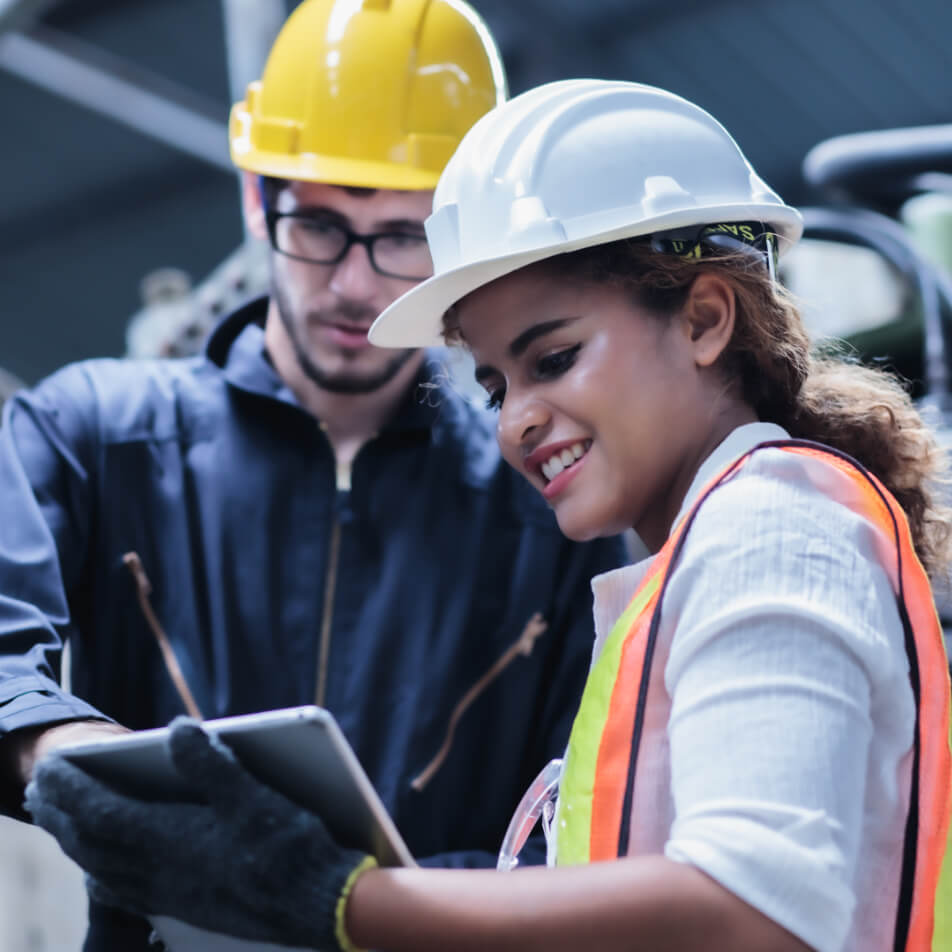 Zwei Arbeiter in Sicherheitsausrüstung, ein Mann mit gelbem Schutzhelm und eine Frau mit weißem Schutzhelm und Sicherheitsweste, schauen in einer Industrieanlage auf ein Tablet.