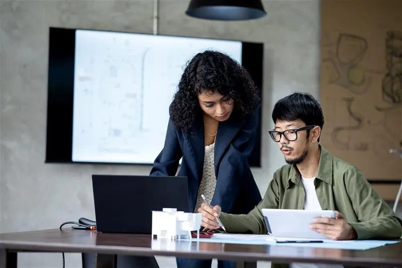 Zwei valantic‑Mitarbeitende arbeiten gemeinsam an einer CX‑Strategy: Frau steht neben einem sitzenden Kollegen, beide betrachten Dokumente am Schreibtisch mit Laptop.