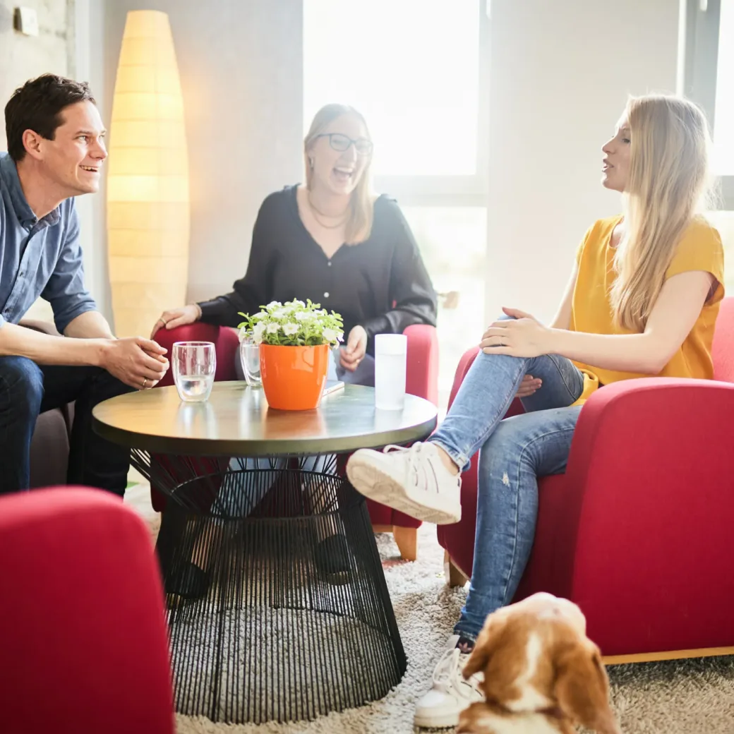 Foto von drei Kolleg*innen, die in einer Lounge-Area Pause machen.
