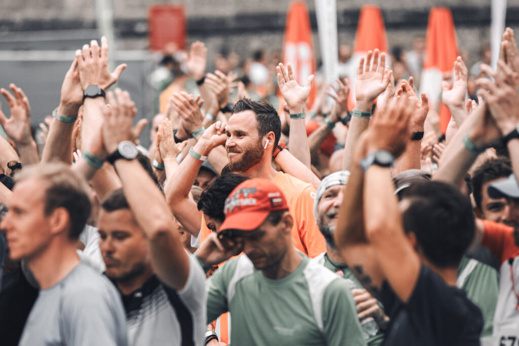Eine große Gruppe Läufer*innen beim Salzburger Businesslauf, die mit erhobenen Händen applaudieren. In der Mitte sticht ein lächelnder elements-Mitarbeiter mit Kopfhörern in den Ohren und einem orangen Sportshirt hervor.