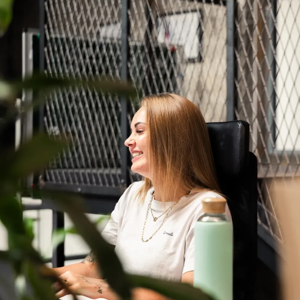 Eine Frau sitzt im Büro am Schreibtisch, lächelt und arbeitet am Computer.