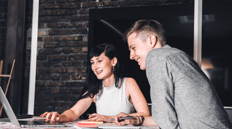 Zwei Personen sitzen an einem Tisch in einem Büro, schauen auf einen Laptop-Bildschirm und lächeln.