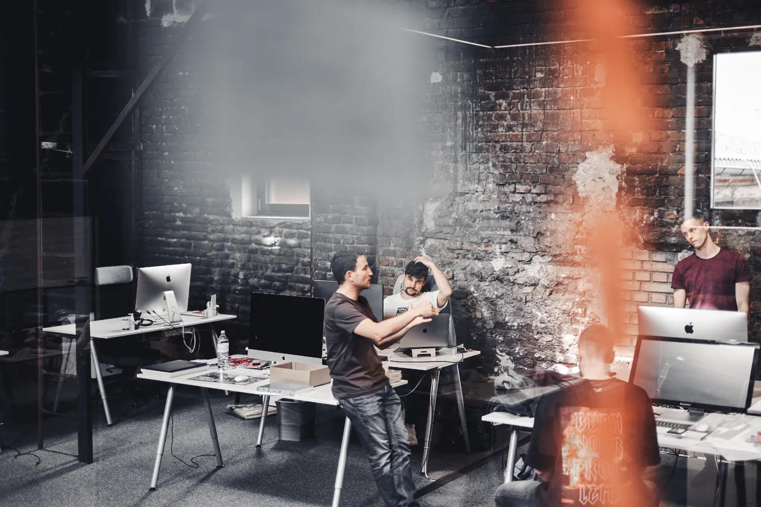 Kreative Arbeitsatmosphäre in einem modernen Büro mit Industrial Design: Mehrere Personen arbeiten und diskutieren gemeinsam an digitalen Projekten zwischen iMac-Arbeitsplätzen und offenen Arbeitsbereichen. Die Szene vermittelt kollaboratives Arbeiten, agile Teamprozesse und die Entwicklung digitaler Lösungen in einem dynamischen Umfeld.