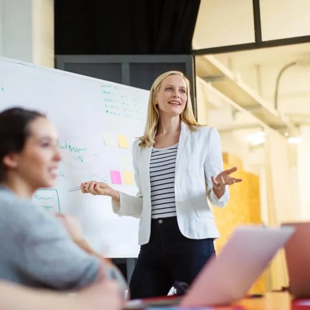 Eine Frau steht vor einem Whiteboard und präsentiert drei sitzenden Kollegen in einem Büro, auf dem Tisch liegen Laptops und Notizen.