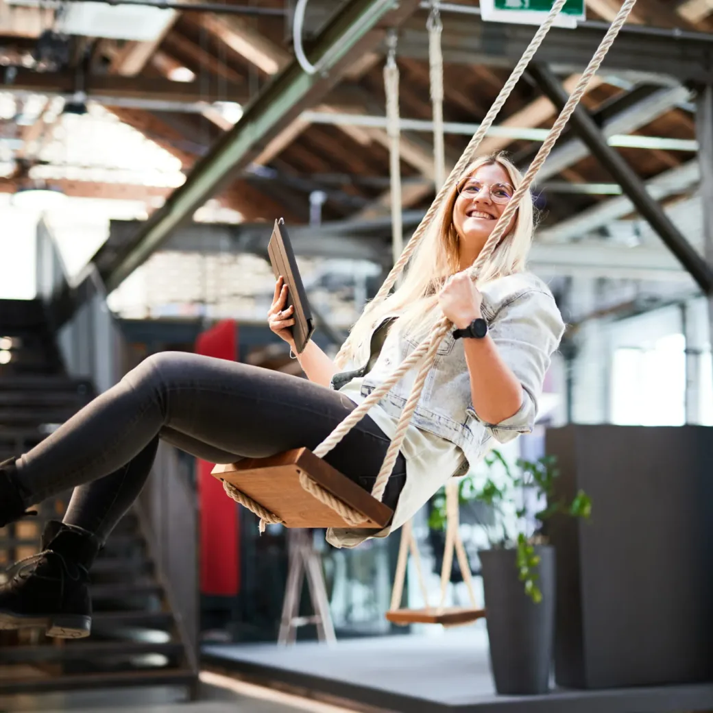 Picture of a young woman with a tablet in her hand, swinging on a swing set in her office.
