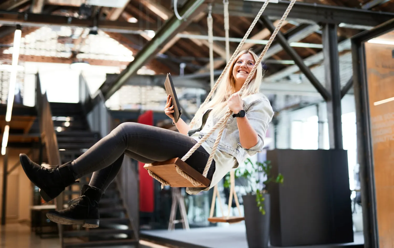 Picture of a young woman with a tablet in her hand, swinging on a swing set in her office.