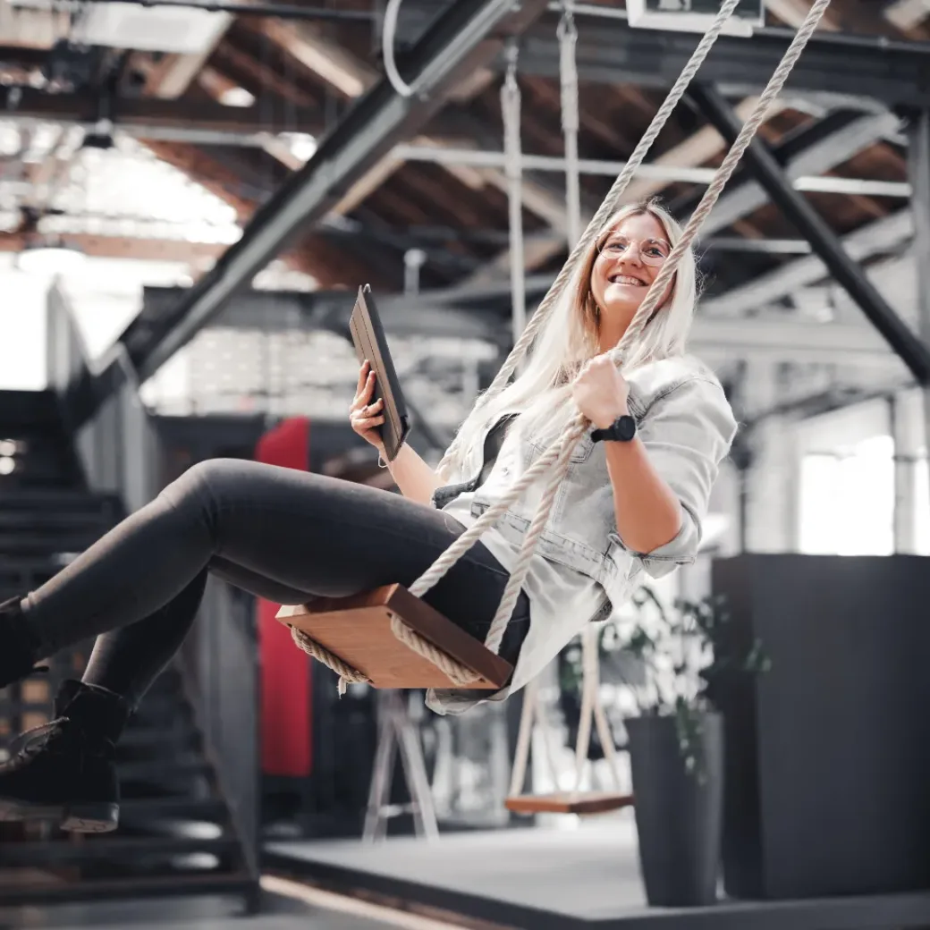 Eine Person sitzt in einem modernen Büroraum im Industriestil auf einer Schaukel, lächelt und hält ein Tablet.