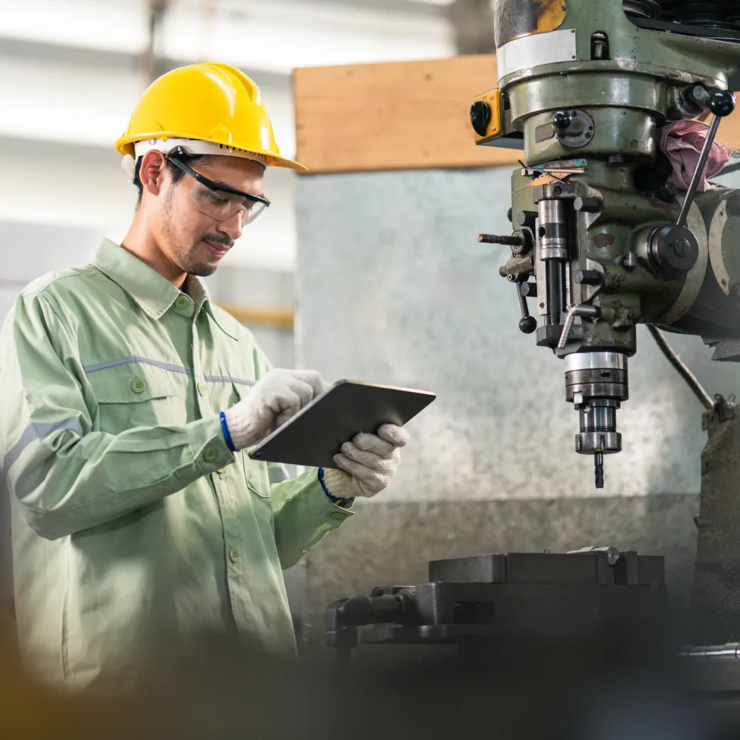 Ein Arbeiter in Schutzkleidung und gelbem Helm hält ein Tablet in den Händen und steht neben einer großen Industriemaschine in einer Fabrikumgebung.