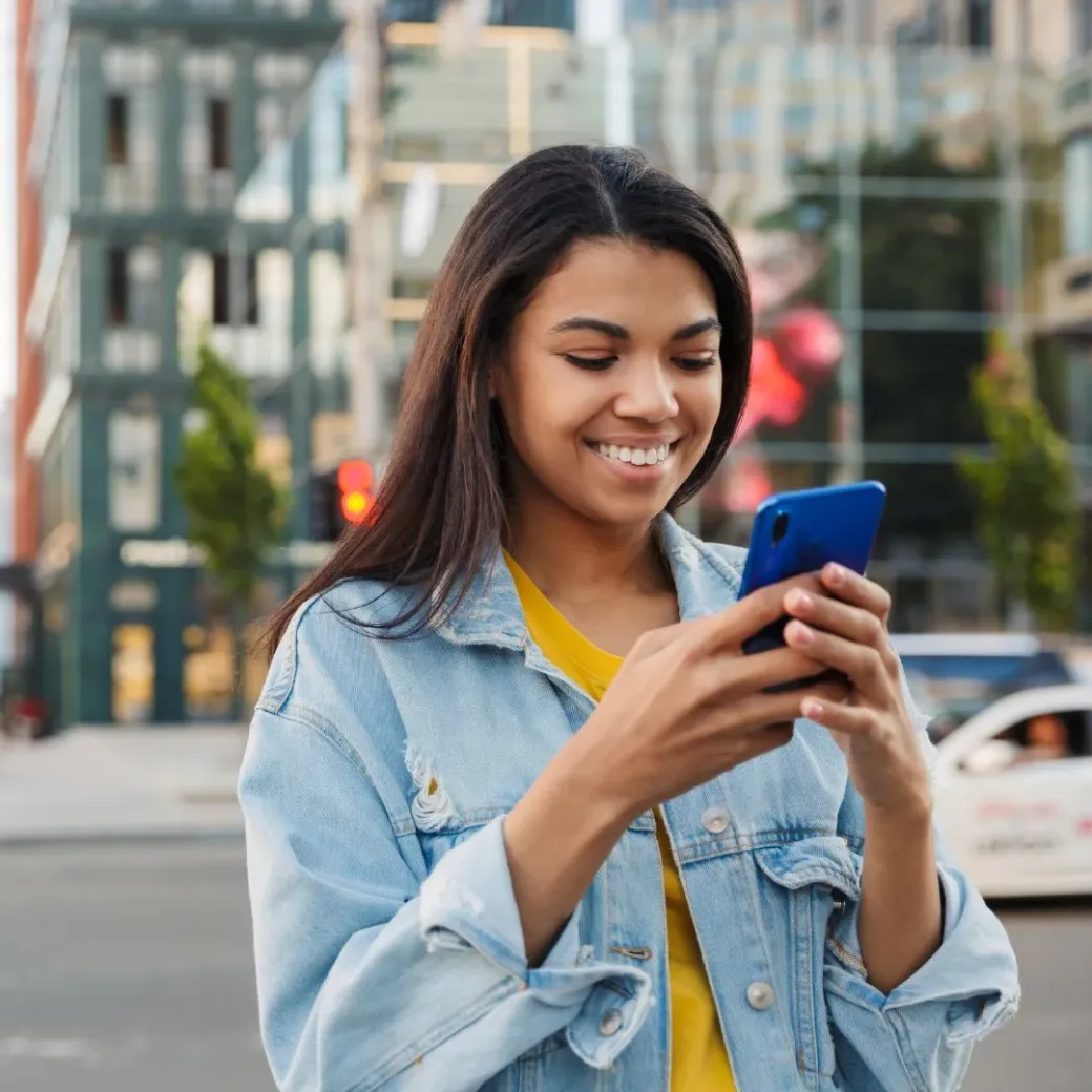 A young woman in a denim jacket is standing outside in a city, smiling as she looks at her smartphone.