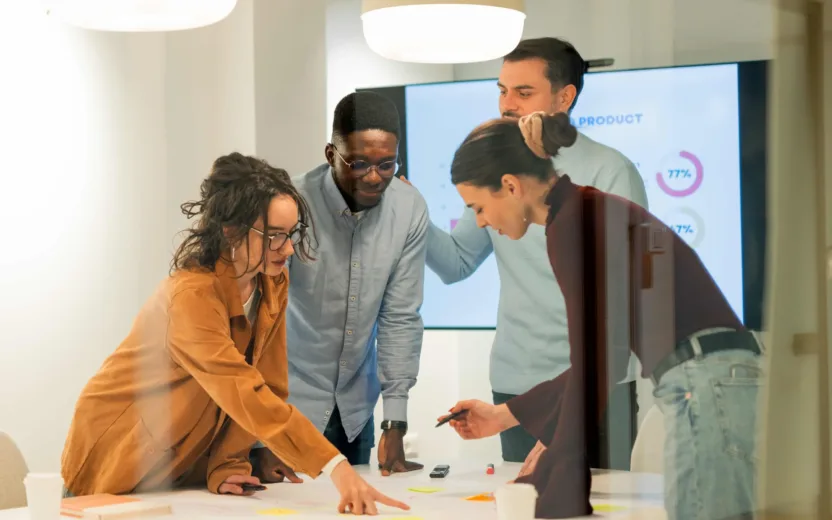 Four people work together in a meeting room at a table with post-its; a screen shows diagrams in the background.