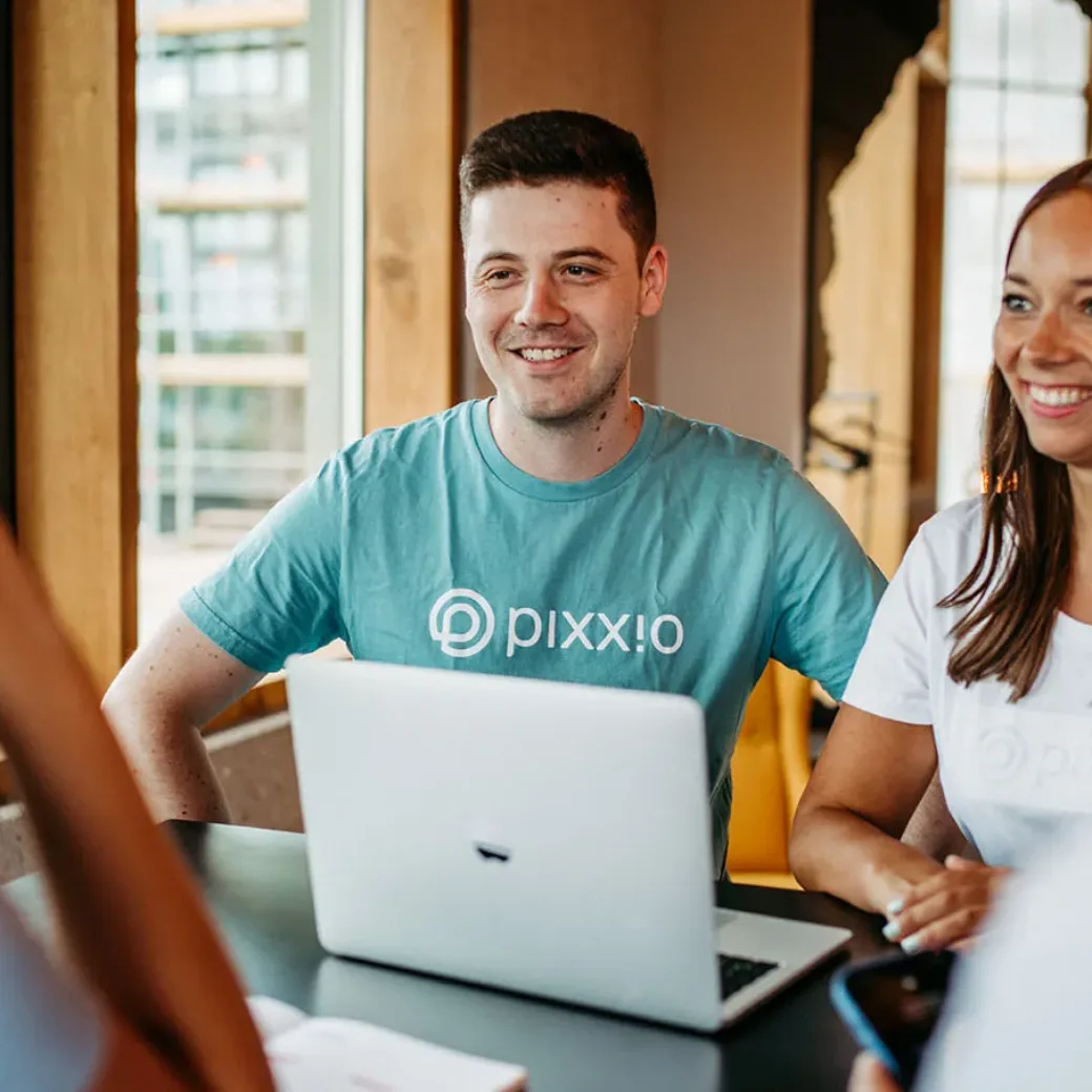 Zwei Personen in Pixx.io-Shirts sitzen an einem Tisch mit einem Laptop, diskutieren über DAM-Lösungen und lächeln mit zwei weiteren Personen in einer modernen, hellen Büroumgebung.