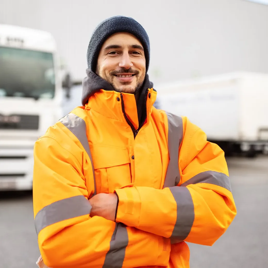 Portrait of a male commercial docker standing outdoors with arms crossed. Shipping yard worker with truck in background.
