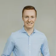 Ein Mann mit kurzen rötlich-braunen Haaren, der ein hellblaues Button-up-Hemd trägt, steht lächelnd vor einem schlichten hellen Hintergrund.