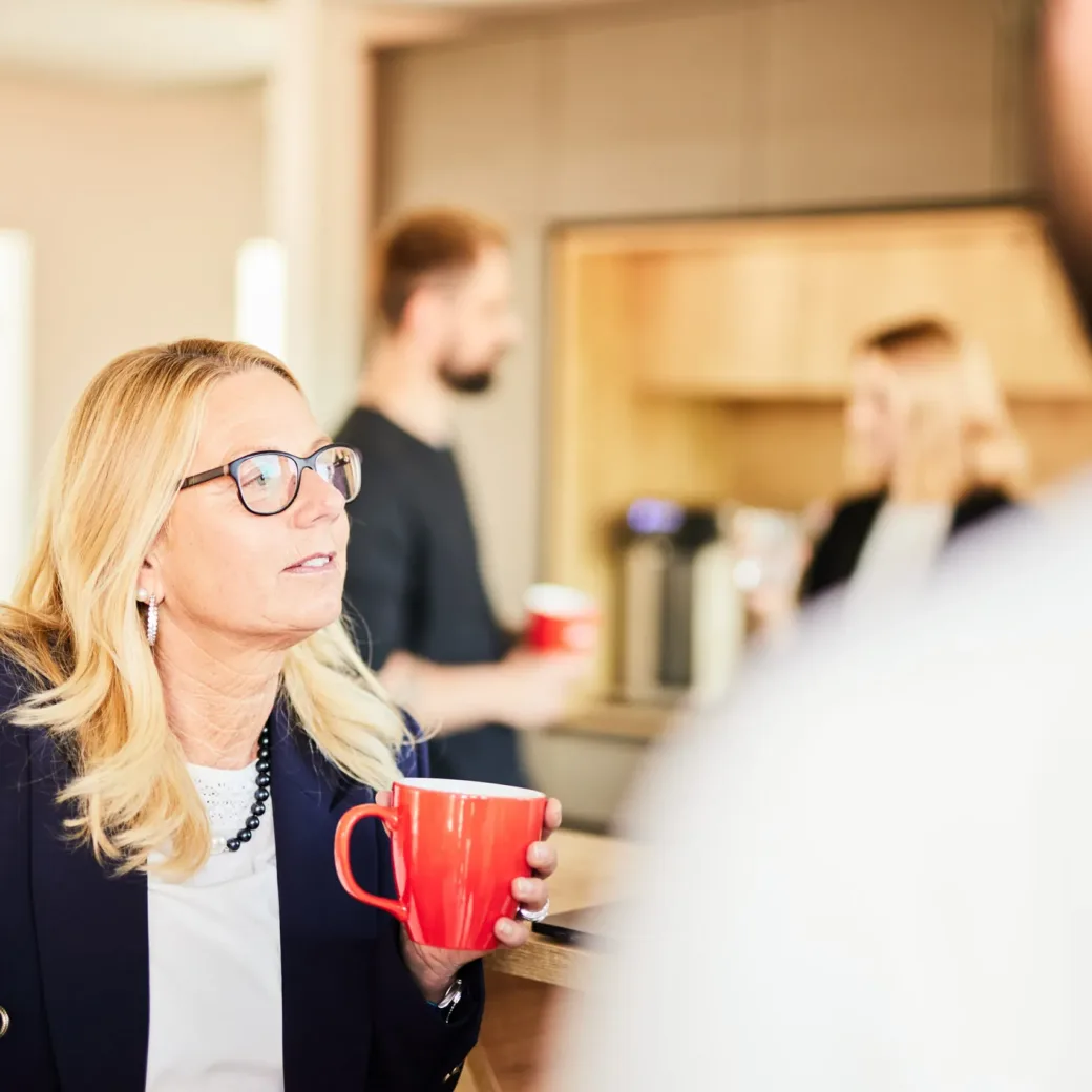 Photo of a blonde middle-aged woman having a conversation with her colleagues at the coffee bar with a valantic cup.
