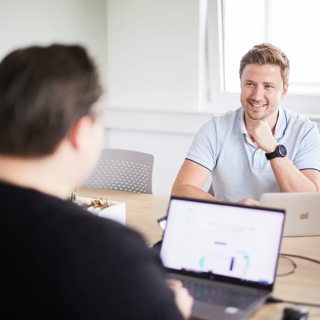 Photo of two colleagues working on their laptops in an office and talking to each other.