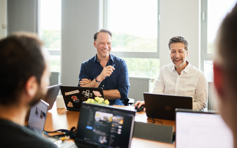 Photo of two men sitting in a meeting with their laptops.