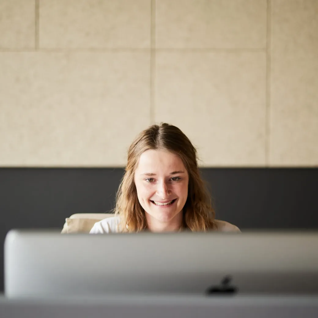 Foto von einer jungen, lächelnden Frau, die am PC arbeitet.