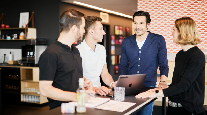 Foto von drei Kolleg*innen die an einem Steharbeitsplatz eine Besprechung abhalten.