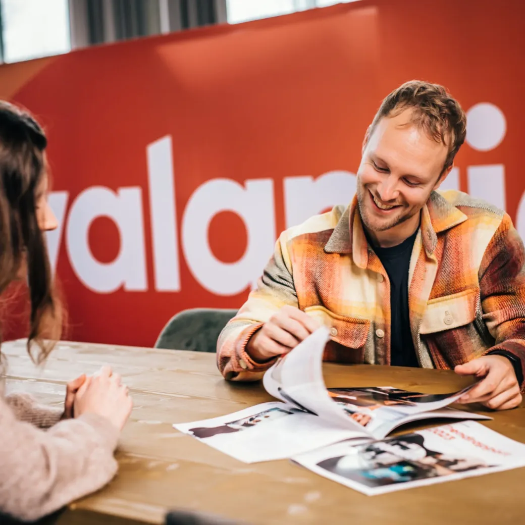 Twee mensen zitten aan een tafel, de een laat de ander een tijdschrift zien. Beiden zijn in gesprek, met een rode muur en een gedeeltelijke tekst 