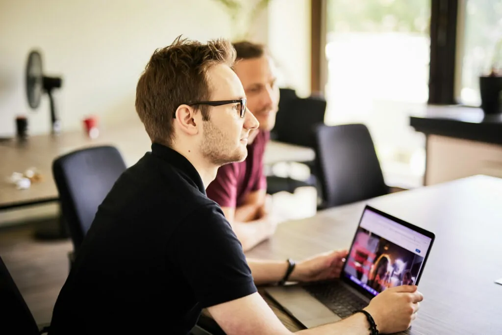 Foto von einem jungen Mann mit Brille, der an seinem Laptop sitzt und zu einem Kollegen nach vorne sieht.