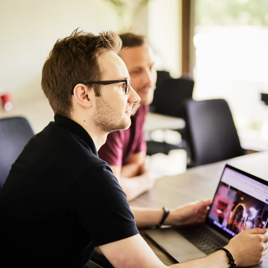 Photo of a young man with glasses sitting at his laptop and looking at a colleague.