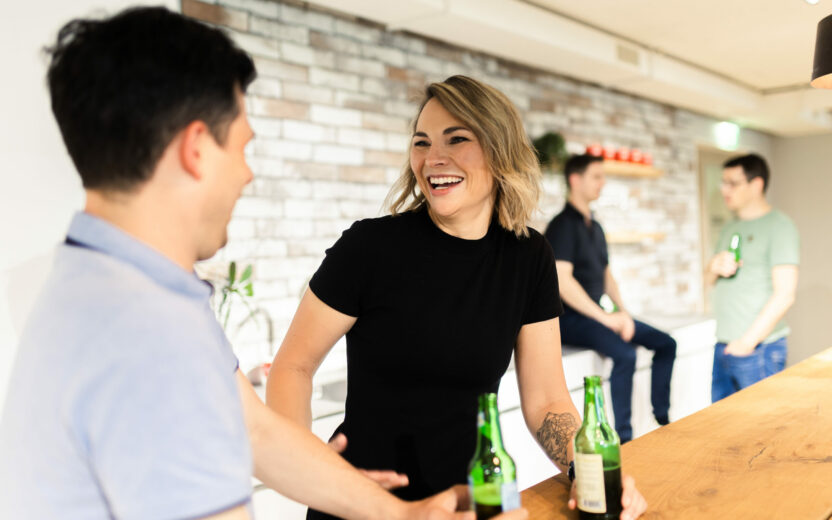 Two people are smiling and holding beer bottles while standing at a wooden counter. Others are in the background, also holding drinks and engaging in conversation.
