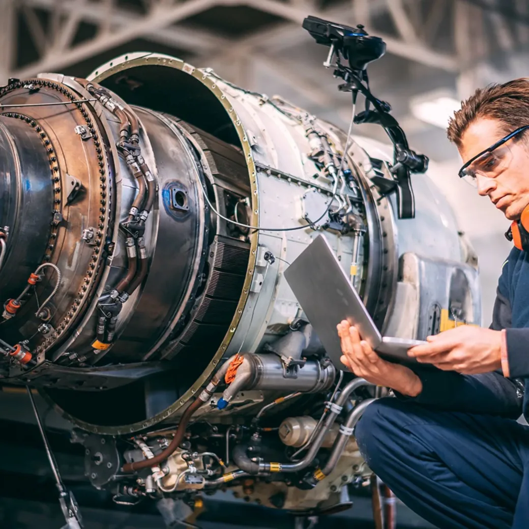 Aircraft engineer in a hangar using a laptop while repairing and maintaining an airplane jet engine