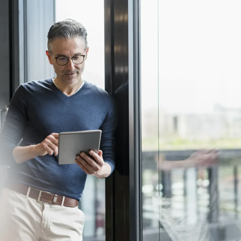 Ein Mann mit Brille steht an einem Fenster, trägt einen dunklen Pullover und eine helle Hose und prüft mit einem Tablet-Gerät Updates zur Cybersicherheit.