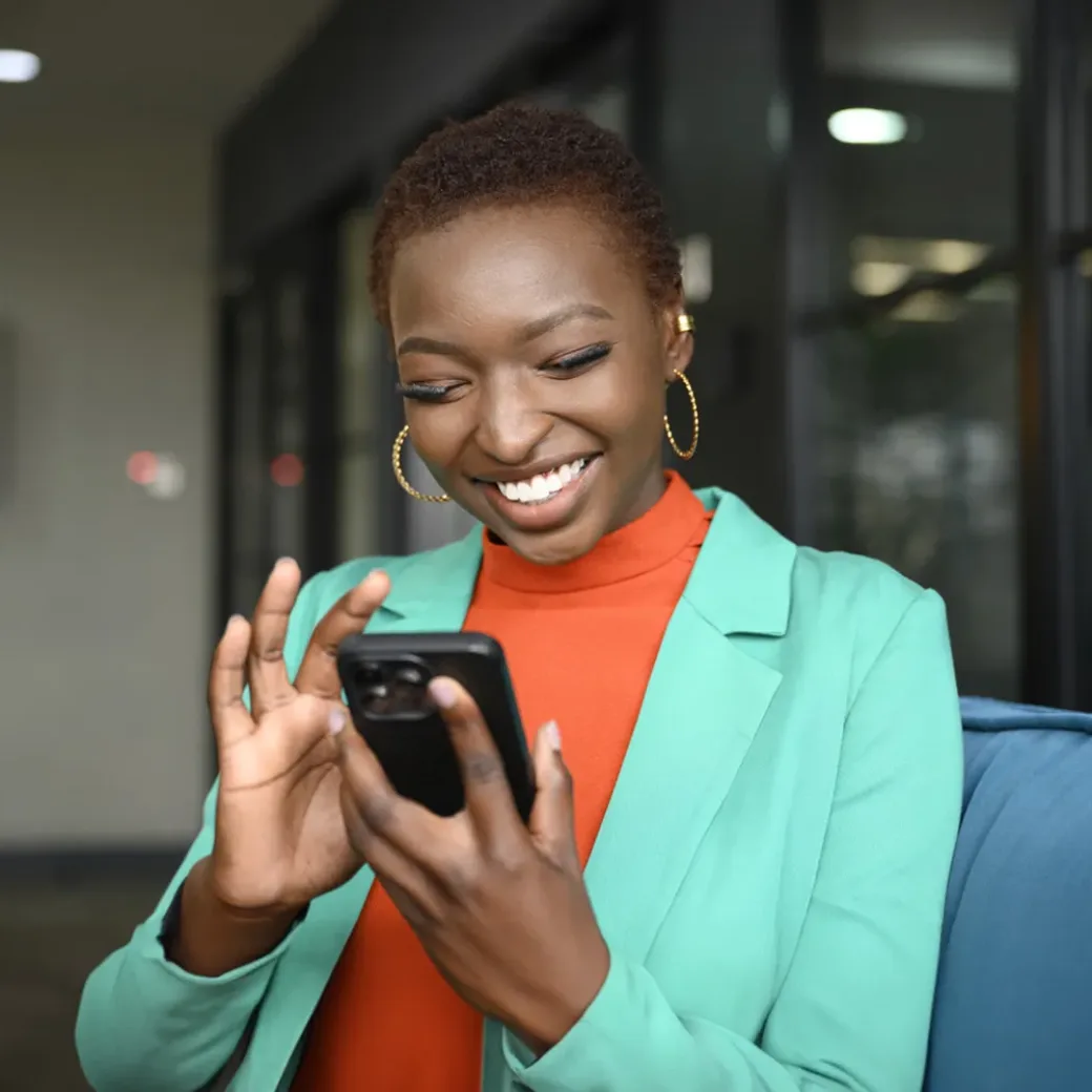 Waist-up view of shorthaired woman wearing hoop earrings, teal jacket over orange top, and smiling while scrolling portable device in modern office.