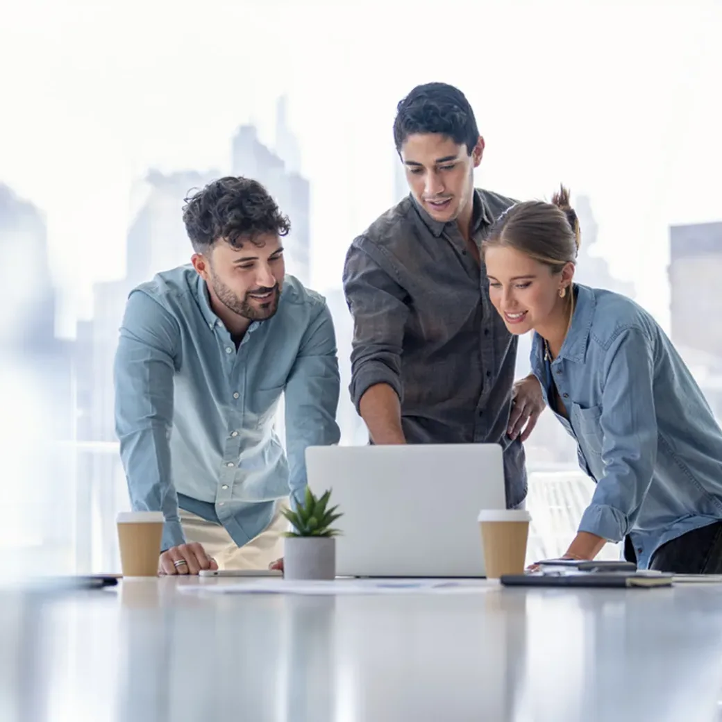 Business team working on a laptop computer. Three people are wearing casual clothing. They are standing in a board room. Multi ethnic group with Cauc and o men and women