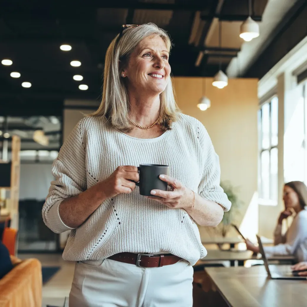 Thoughtful businesswoman standing in a co-working space. Mature businesswoman smiling cheerfully while holding a cup of coffee. Experienced entrepreneur contemplating new ideas during her coffee break.