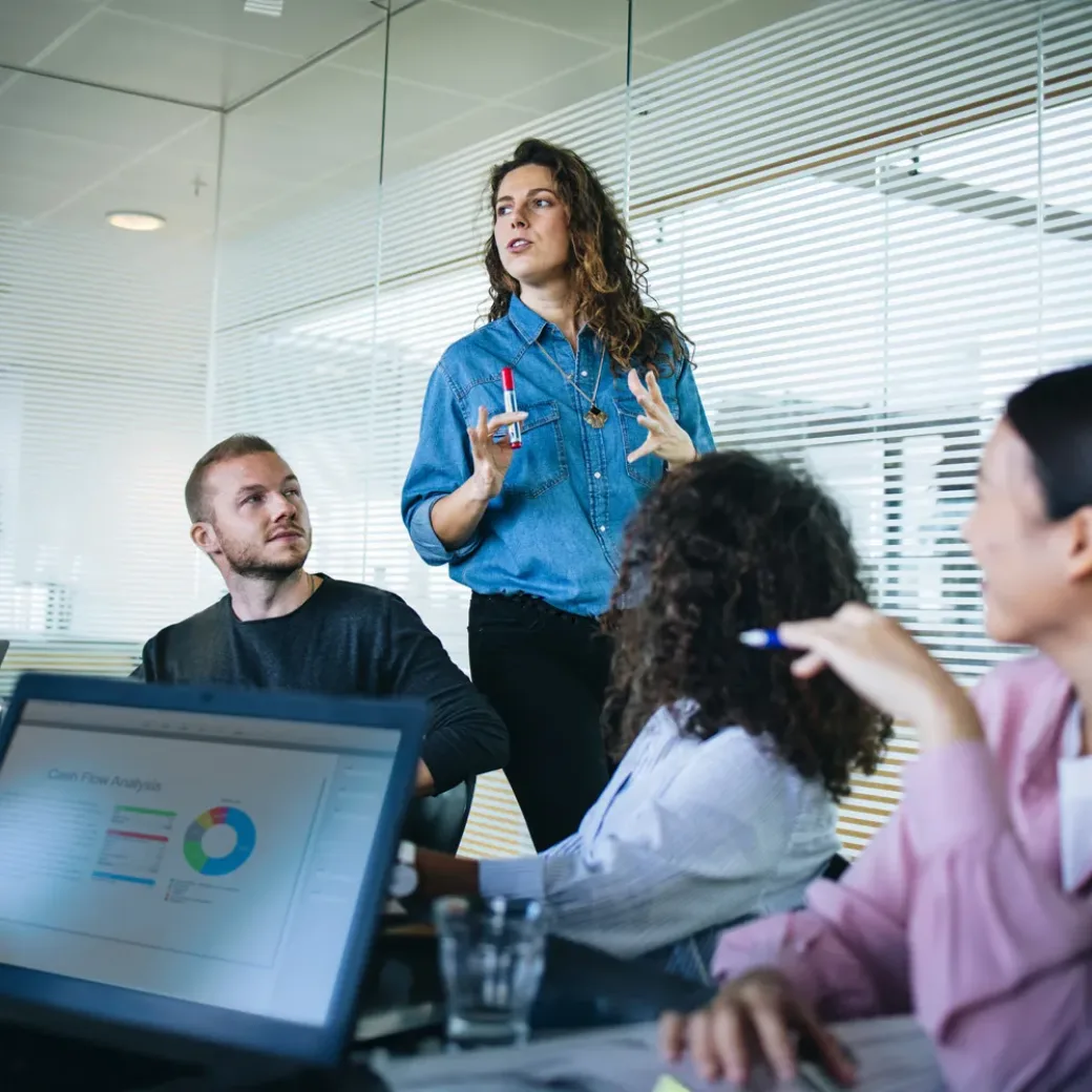 Businesswoman walking in office boardroom and sharing a strategy with team sitting at table. Female manager explaining new business plan with coworkers sitting at conference table in office.