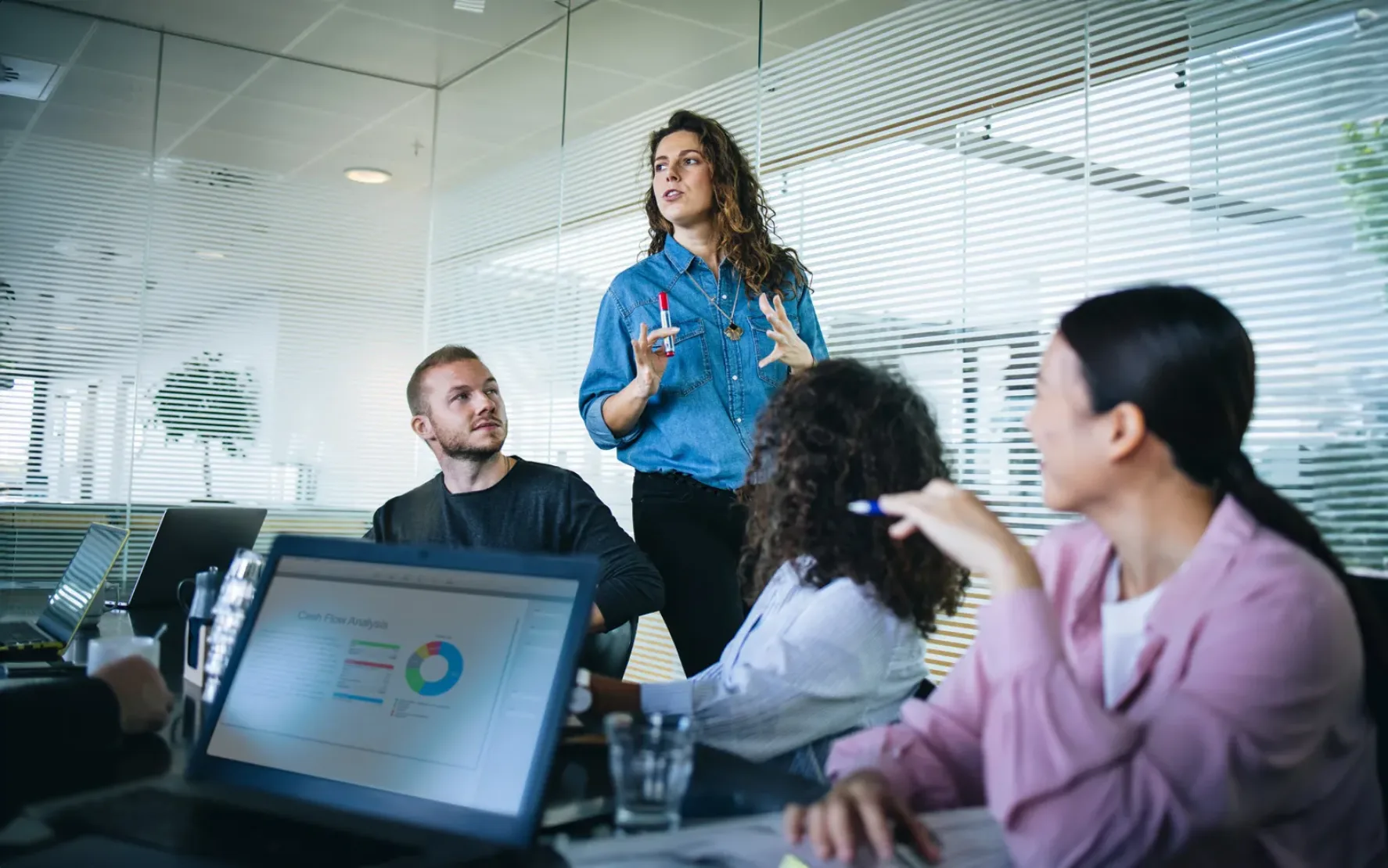 Businesswoman walking in office boardroom and sharing a strategy with team sitting at table. Female manager explaining new business plan with coworkers sitting at conference table in office.