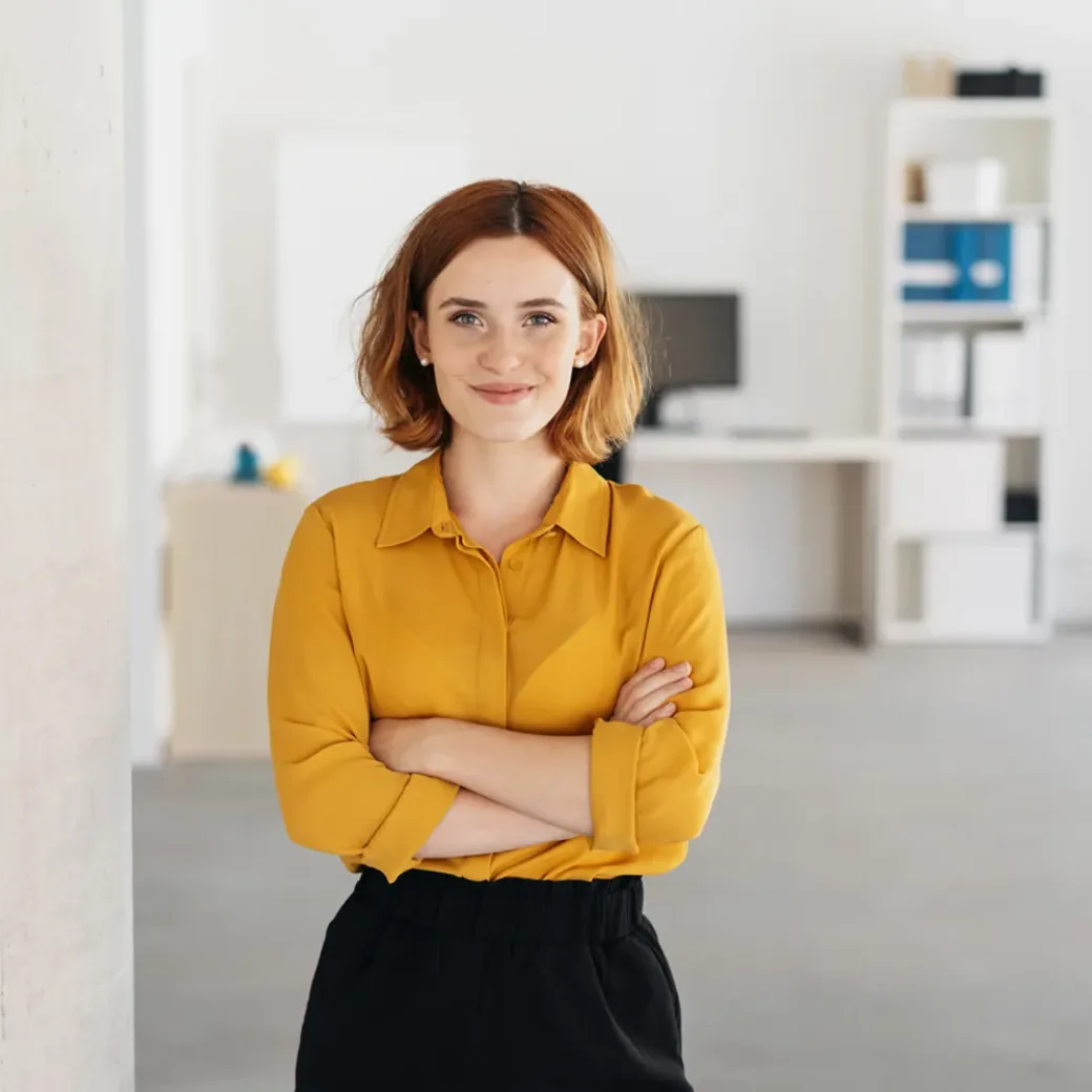 Happy relaxed confident young businesswoman standing with folded arms in a spacious office looking at the camera with a warm friendly smile