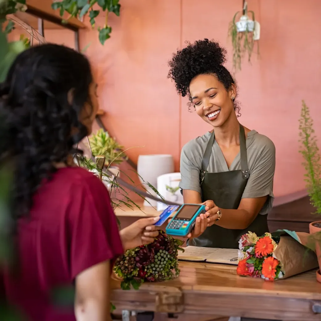 Smiling and friendly florist holding card reader machine at counter with customer paying with credit card.