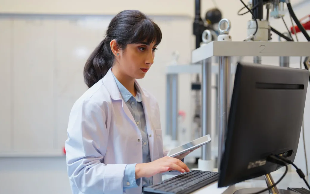 Eine Frau in einem Laborkittel benutzt eine Tastatur und ein Tablet, während sie an einem Computer in einer Laborumgebung arbeitet.