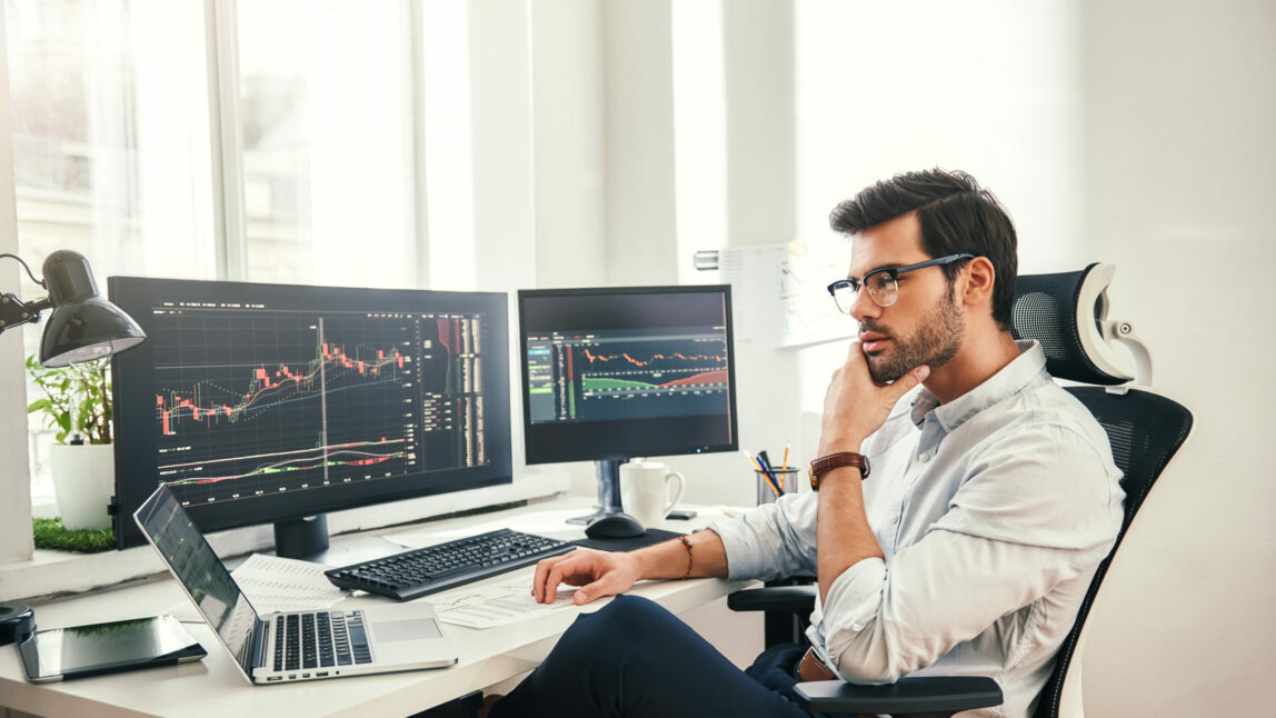 Imagem de um homem sentado em frente a vários ecrãs de compurador com gráficos, setor financeiro