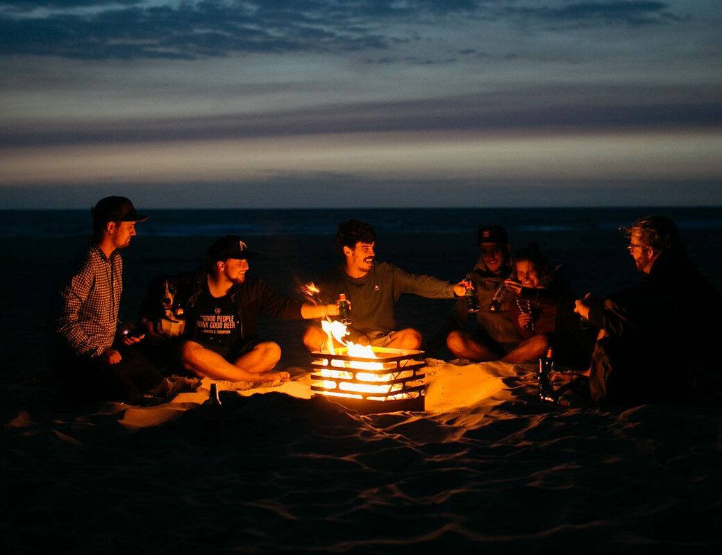 A group of young people sit around a campfire at sunset with the Cube fire basket from höfats.