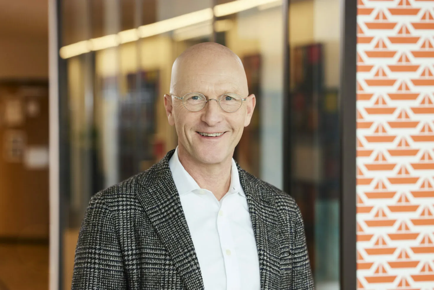 A smiling man with glasses is wearing a checkered blazer and white shirt. He is standing indoors with glass panels and patterned wall in the background.