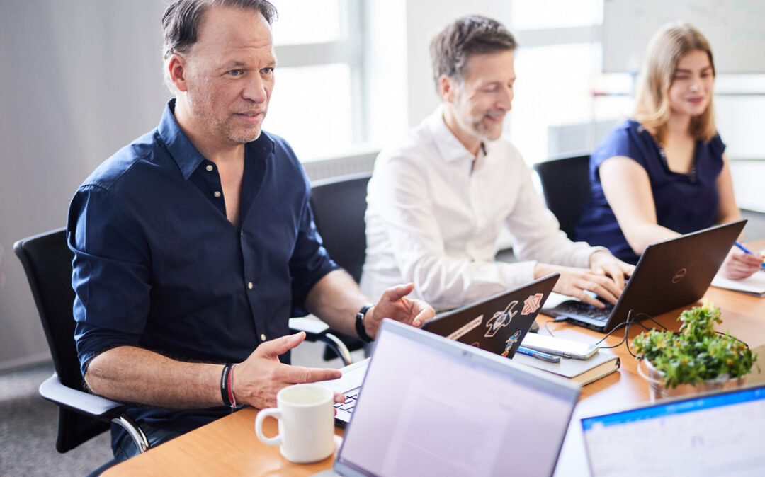 Drei Personen sitzen mit Laptops an einem Konferenztisch und diskutieren.