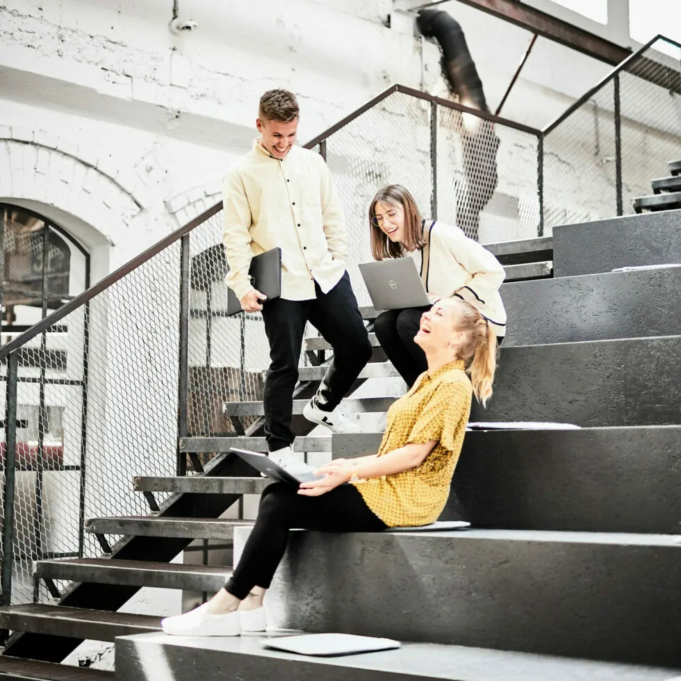 Bild von drei Kolleg*innen, die auf großen Stufen sitzen oder stehen und ausgelassen lachen.
