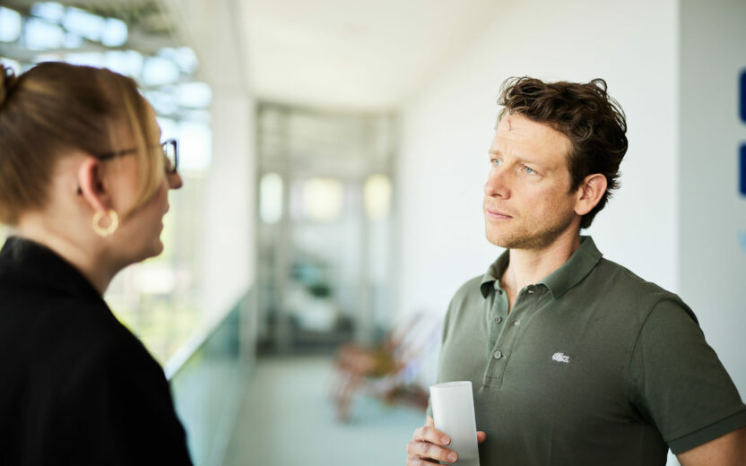 A man holding a cup is engaged in a conversation with a woman wearing glasses. They are standing in a brightly lit indoor space with a glass railing visible in the background.
