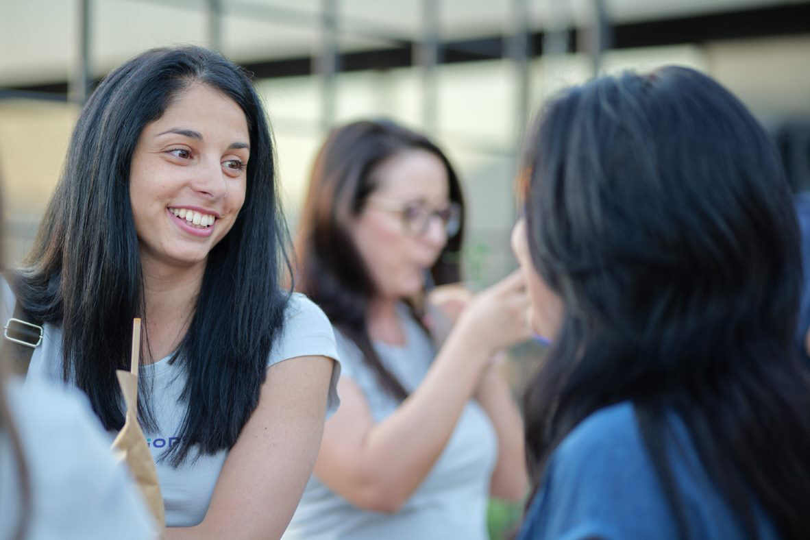Two women, one with long dark hair smiling, the other with long dark hair facing away, are engaged in conversation outdoors. A third woman in the background is looking down while holding a drink.