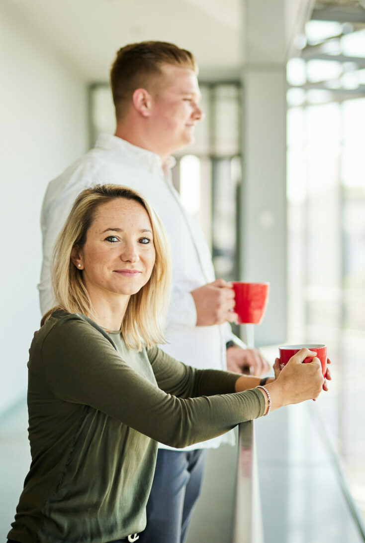 Ein Mann und eine Frau, die jeweils mit einer Tasse am Balkongelände ihrer Firma stehen.
