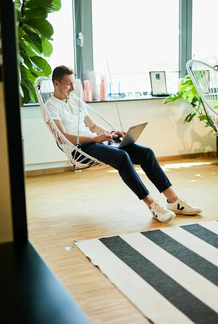 A man sits on a hanging chair in an office, working on a laptop. There are plants, a striped rug, and large windows in the background.