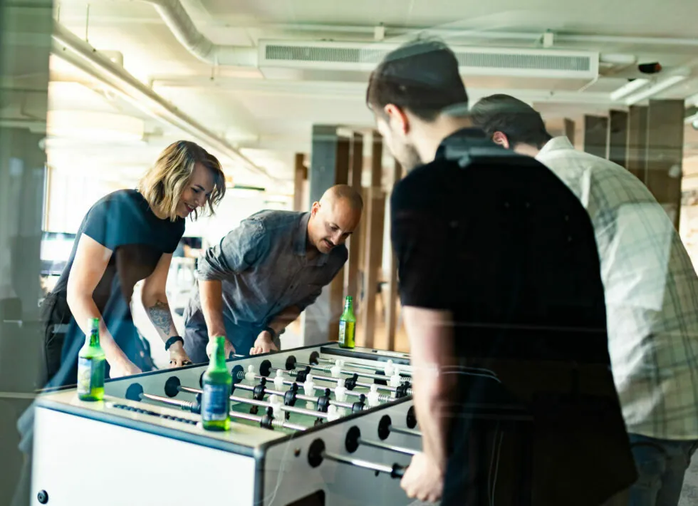 Four people are engaged in a foosball game, with drinks placed on the table's edges in an indoor setting.