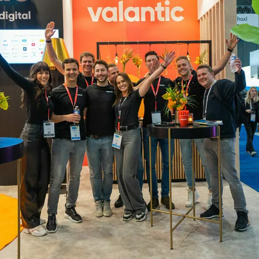 A group of people wearing black shirts, standing in front of an orange "valantic" booth at a convention, are smiling and raising their arms. They are posing for the camera with visible name badges.