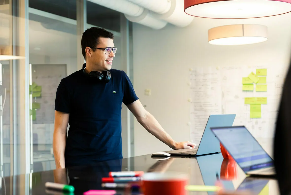 A person stands at a desk, looking at a laptop screen. Various office supplies, a notebook, and a second laptop are also on the desk. Office details and notes are visible in the background.