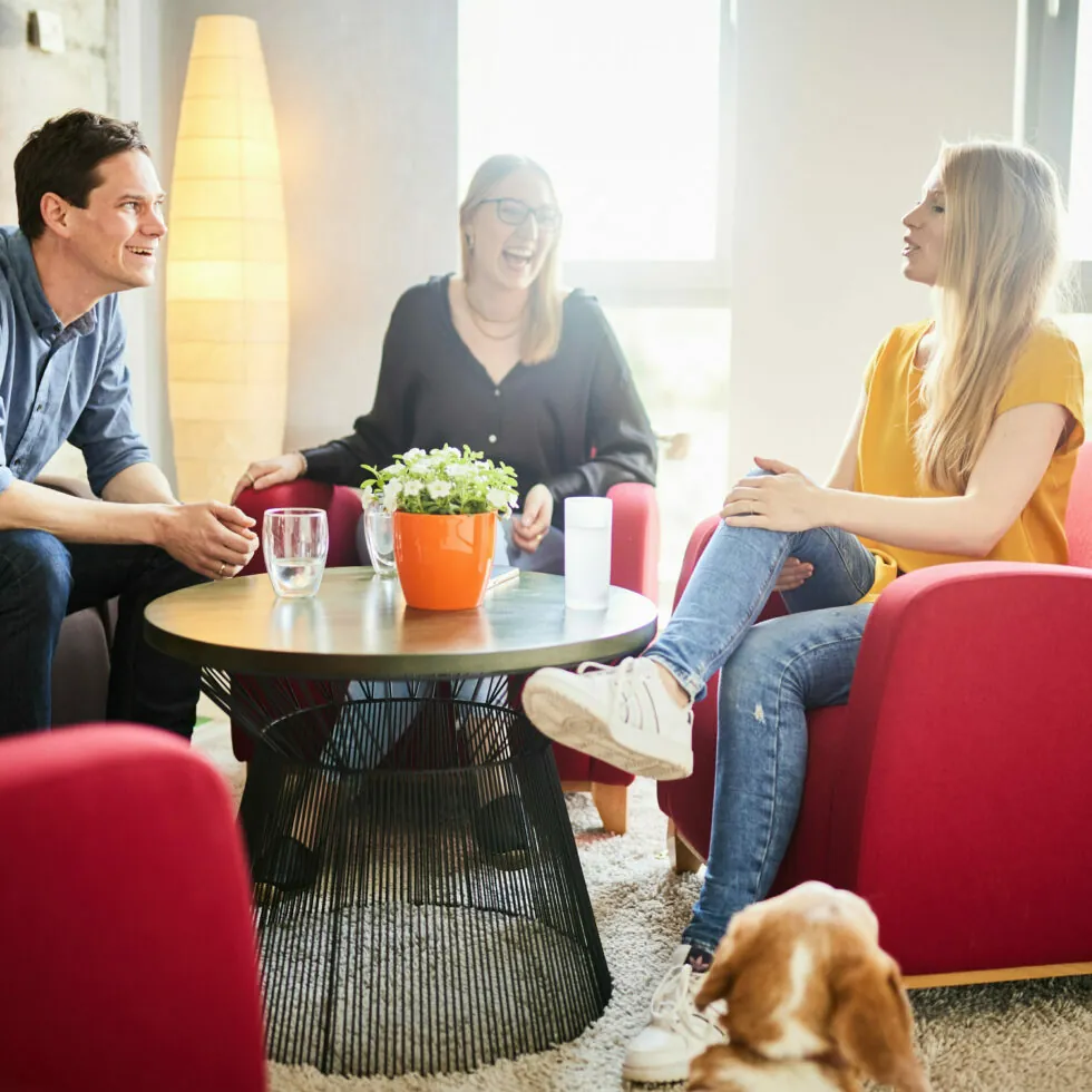 Three people are sitting around a round coffee table, having a conversation and laughing in a brightly lit room. A dog is lying on the floor in the foreground.