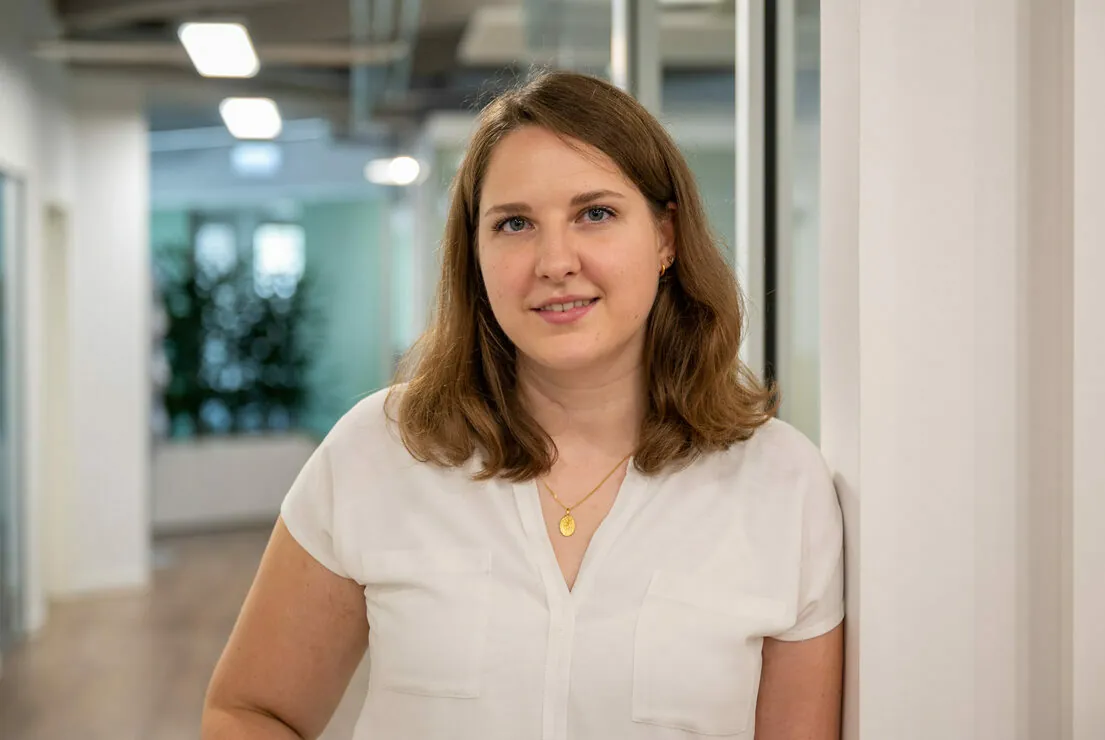 A woman with shoulder-length brown hair, wearing a white blouse and gold necklace, stands in a brightly lit hallway, leaning against a wall.